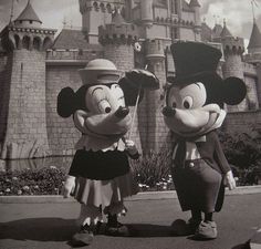 Two Mickey Mouse characters stand in front of a castle, one holding an umbrella, in a 1950s Disneyland costume, capturing nostalgic charm.