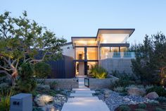 The front of this modern house has a path surrounded by landscaping, that leads to the large, light wood, pivoting front door. #architecture