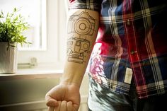 A person with a camera tattoo stands in front of a window, fists clenched, blending photography, art, and self-expression.
