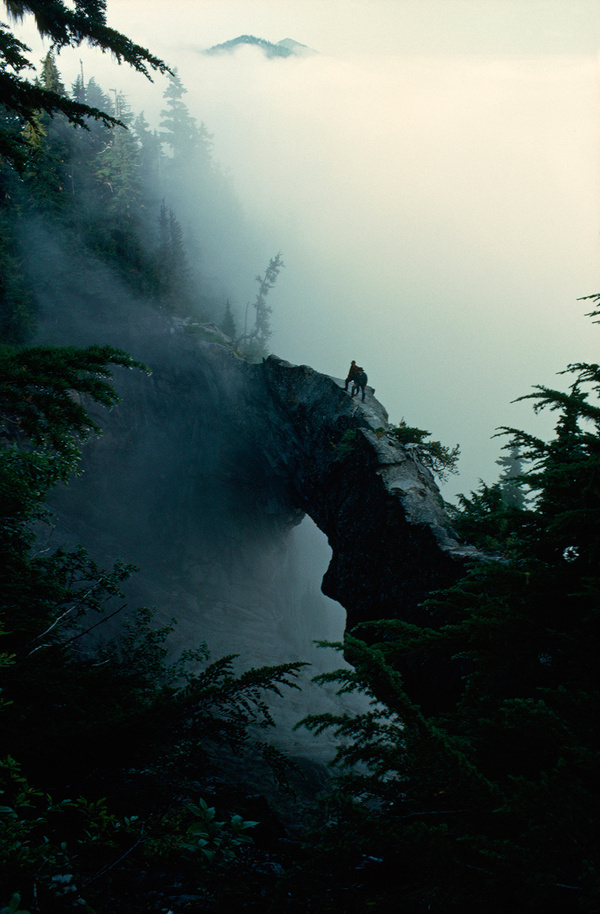 Hikers stand near the top of a natural rock bridge on Mt. Rainier, Washington, May 1963.Photograph by Barry Bishop, National Geographic #fog #1963 #rock #hikers #nat #nature #vintage #mt #film #bridge #geo #rainier