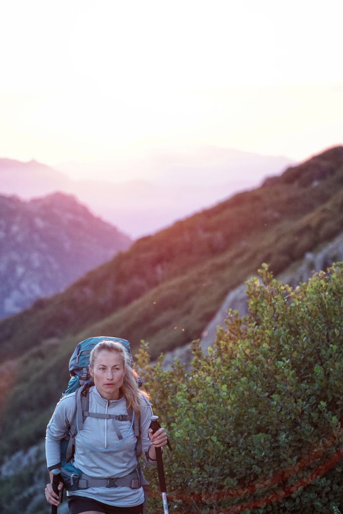 A blonde woman wears a backpack and uses walking poles while hiking near bushes, a sunset over mountains behind her