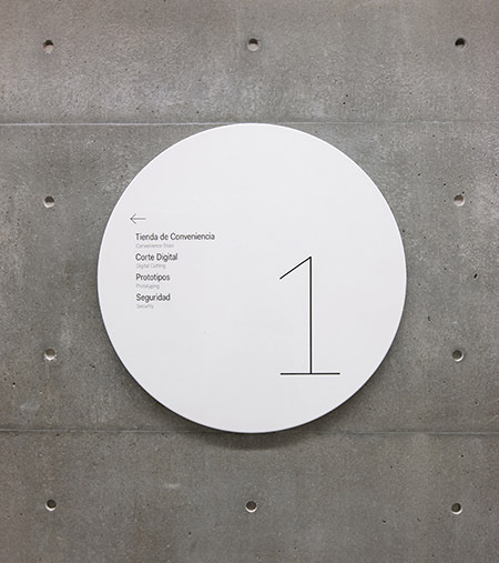 Identification and wayfinding signage appears on shiny metal discs that stand in contrast to the building's textured concrete walls. #textured #concrete #stand #identification #wayfinding #discs #appears #contrast #signage #metal #shiny #walls #buildings