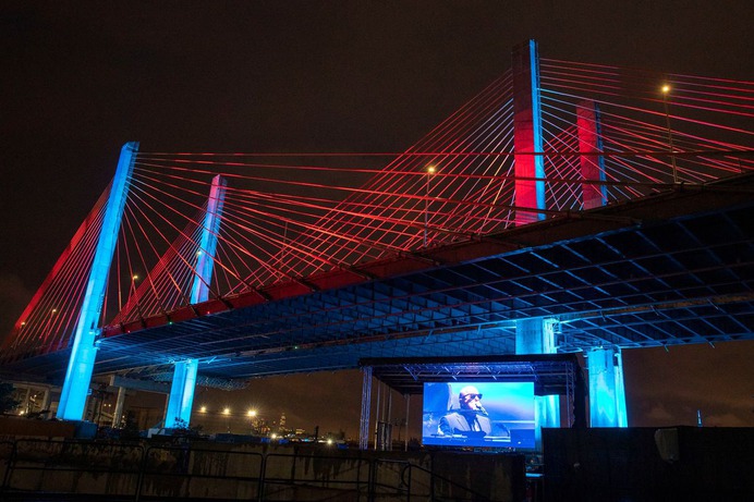 A suspension bridge with steel cables at nighttime. There is a screen under the bridge showing a live performance from a piano player.