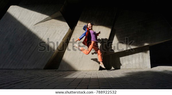 Leaping in flight girl with black hair in blue-orange leather jacket, orange tight pants, colored sneakers. Gray wall with shadow