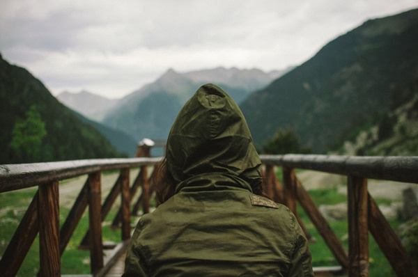 a young woman with her back to us wearing a green hooded jacket standing on a bridge with mountains spread out in front of her #photography