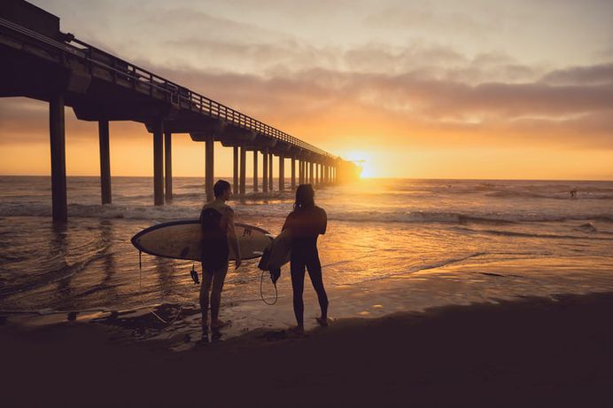 Two surfers holding surfboards looking out at the sunrise and the pier
