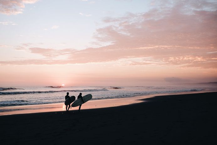 Silhouettes of two people carrying their surfboards on an empty beach during sunset