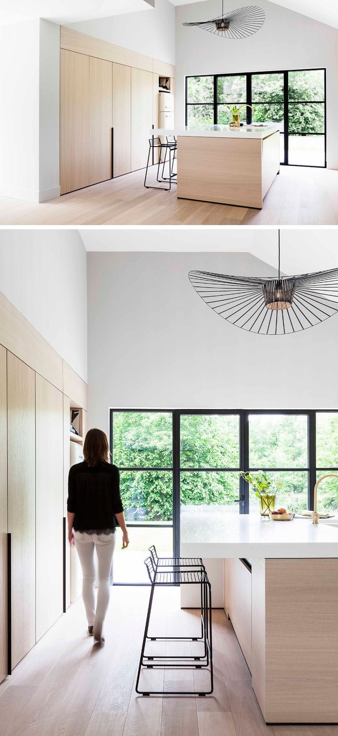 This modern and minimalist light wood and white kitchen features black accents like stools and window frames, a large central island, foldaway cabinet doors, and a pantry with hidden lighting.