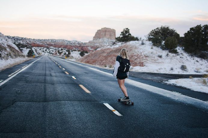 A blonde woman wearing an oversized jacket and backpack skateboarding down a desert road in Page