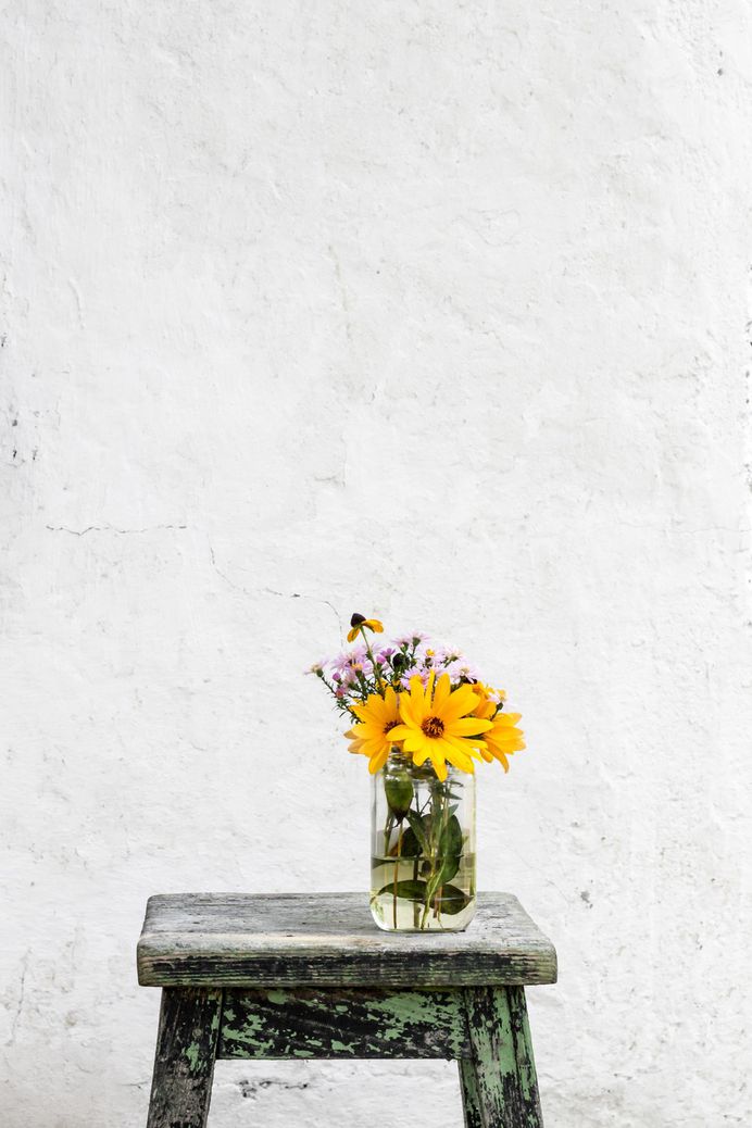 A glass vase with large yellow and small pink flowers on a worn-out stool against an old white wall