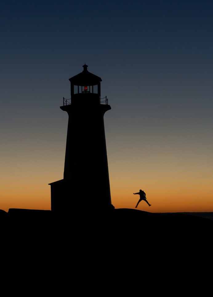 Anonymous traveler jumping with legs apart against lighthouse silhouette and bright sky at night