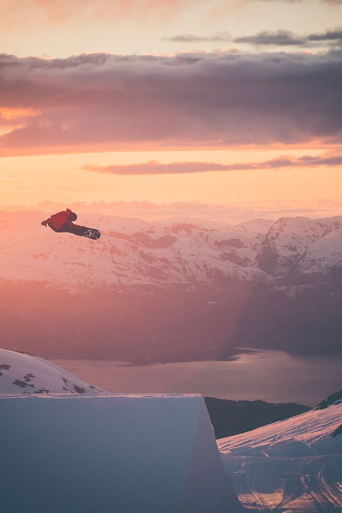 A snowboarder is airborne surrounded by golden skies and snowy mountains